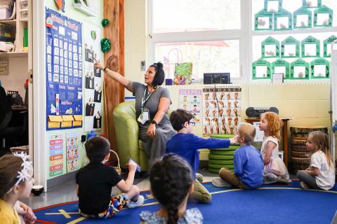 Preschool teacher points to wall calendar during circle time with students watching