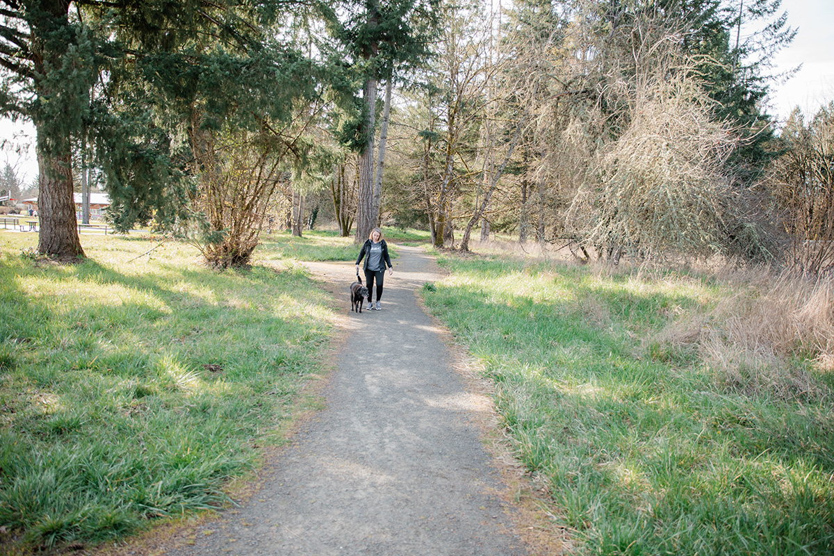 A woman walks with her leashed dog on a gravel path