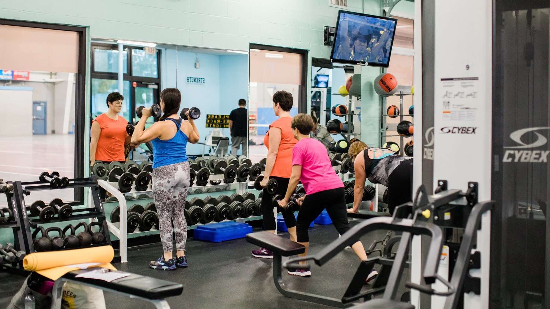 Four people work out in the fitness center lifting weights in front of mirror