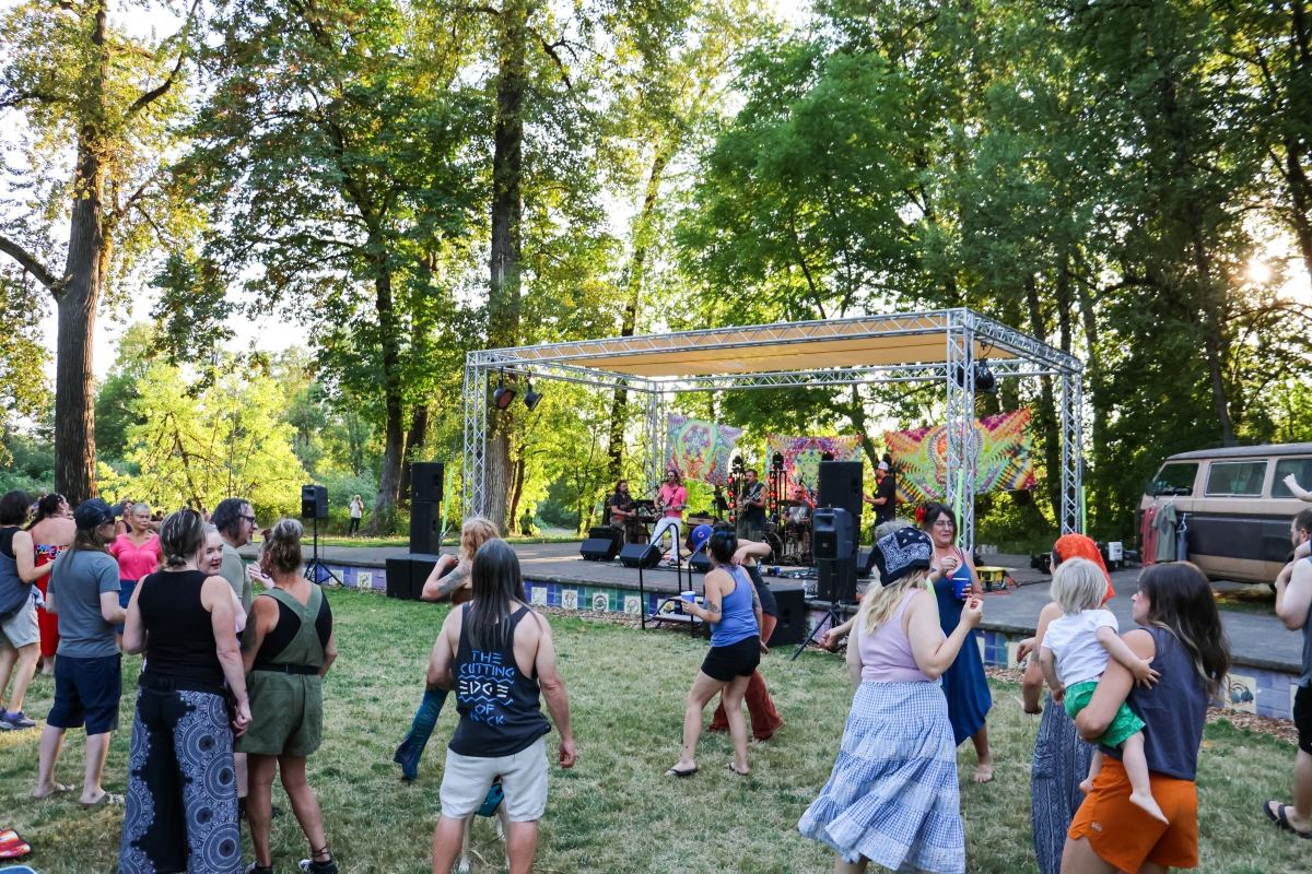 A group of adults and children dancing outside on the grass in front of a stage with a band.