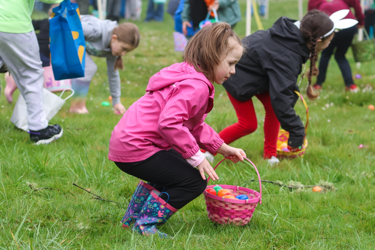 A young child in a pink coat holds a basket filled with colored eggs, while leaning down to pick up another in the green grass.