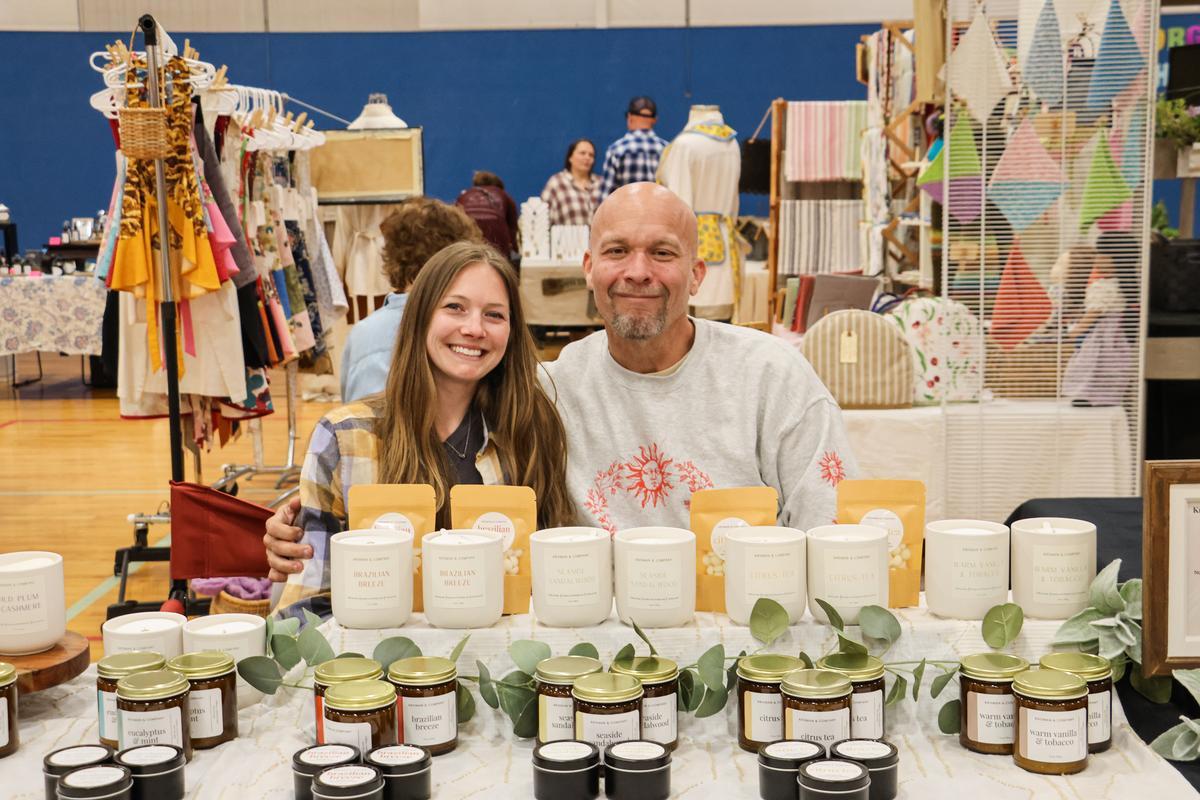 Two adults sit behind a table with plants for sale.
