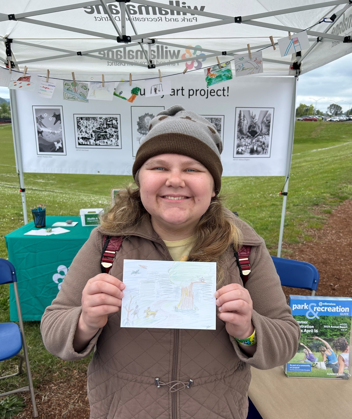 An adult in a brown jacket holds up a hand-drawn postcard of a scene in a park.