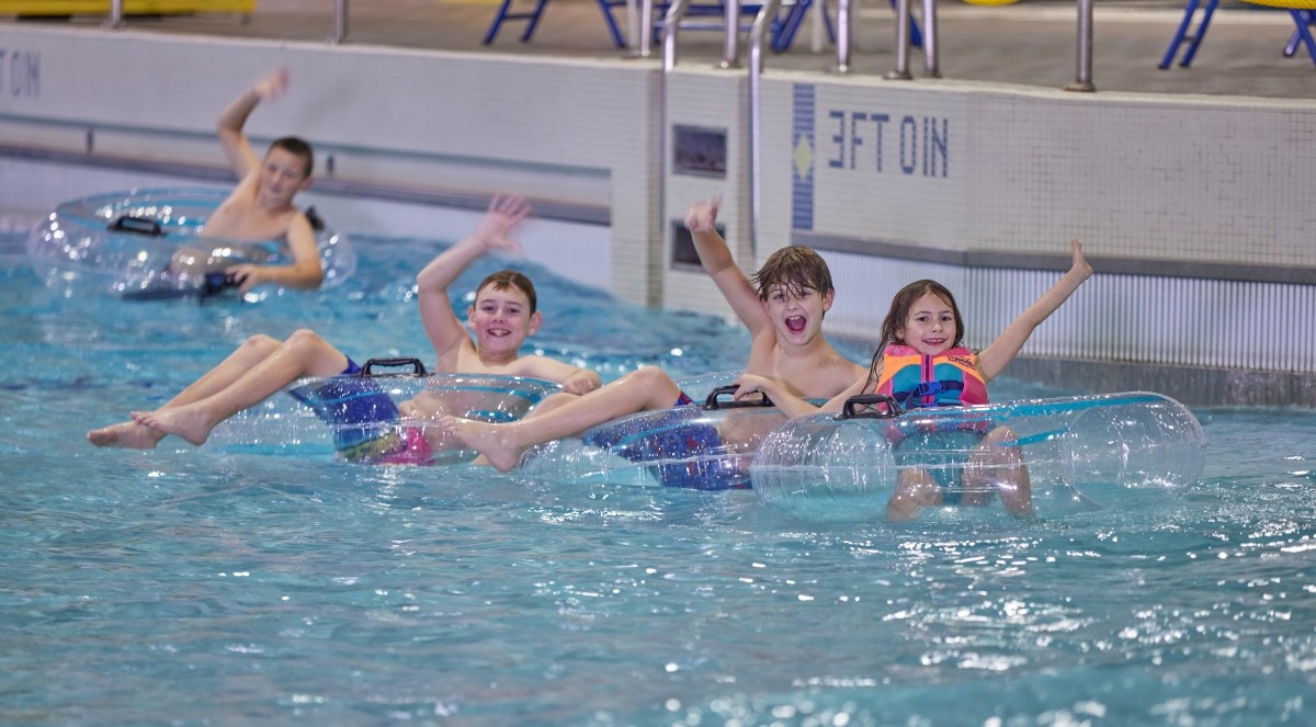 Four kids float on pool water while sitting on top of inflatable tubes. They are smiling and waving.