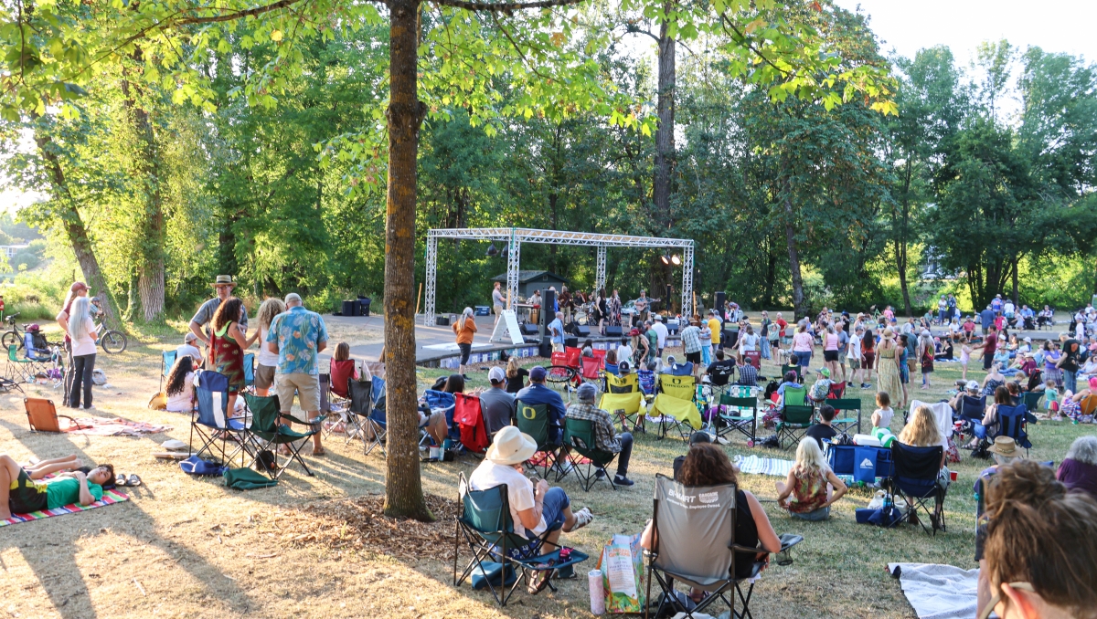 A group of people sit and stand on green grass in front of a large stage with a band playing music.