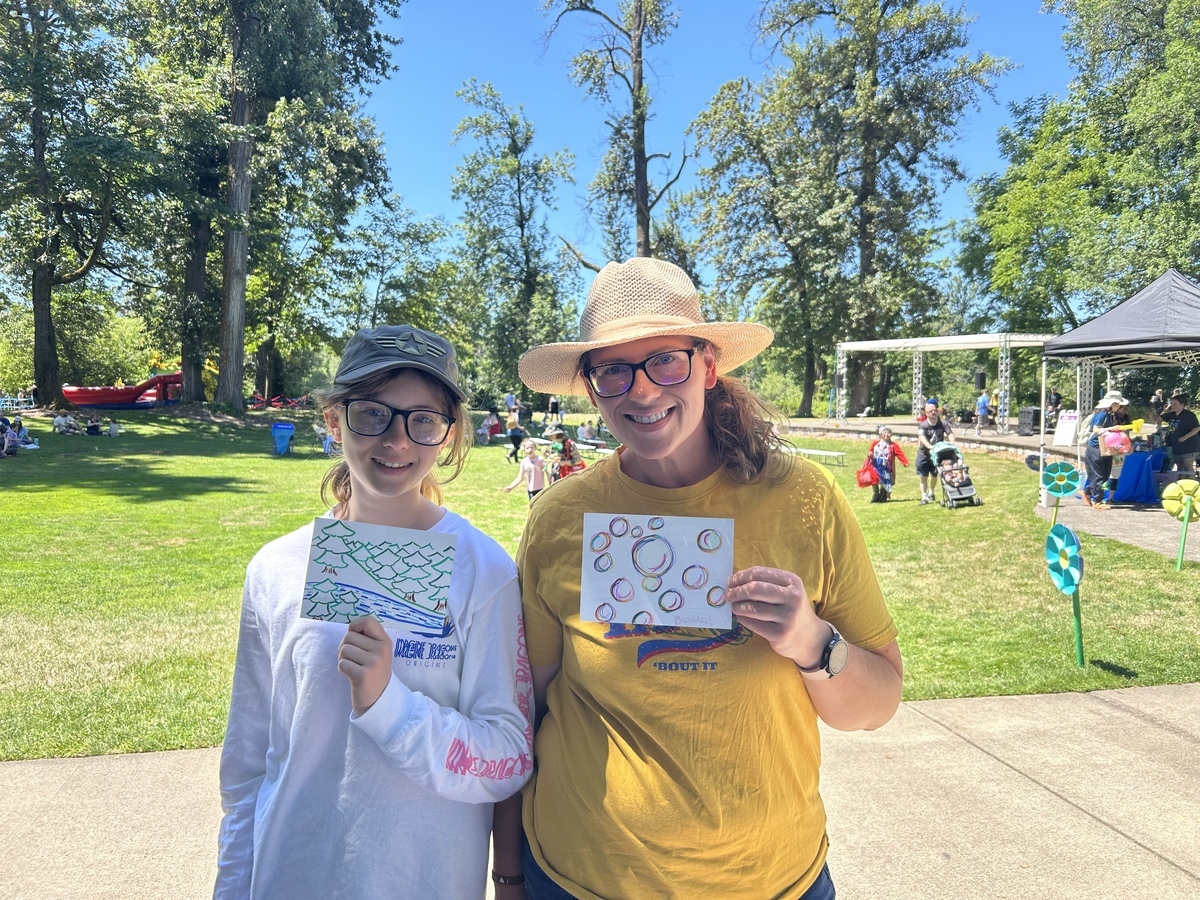 An adult and child stand together holding hand-drawn art outside in a park.