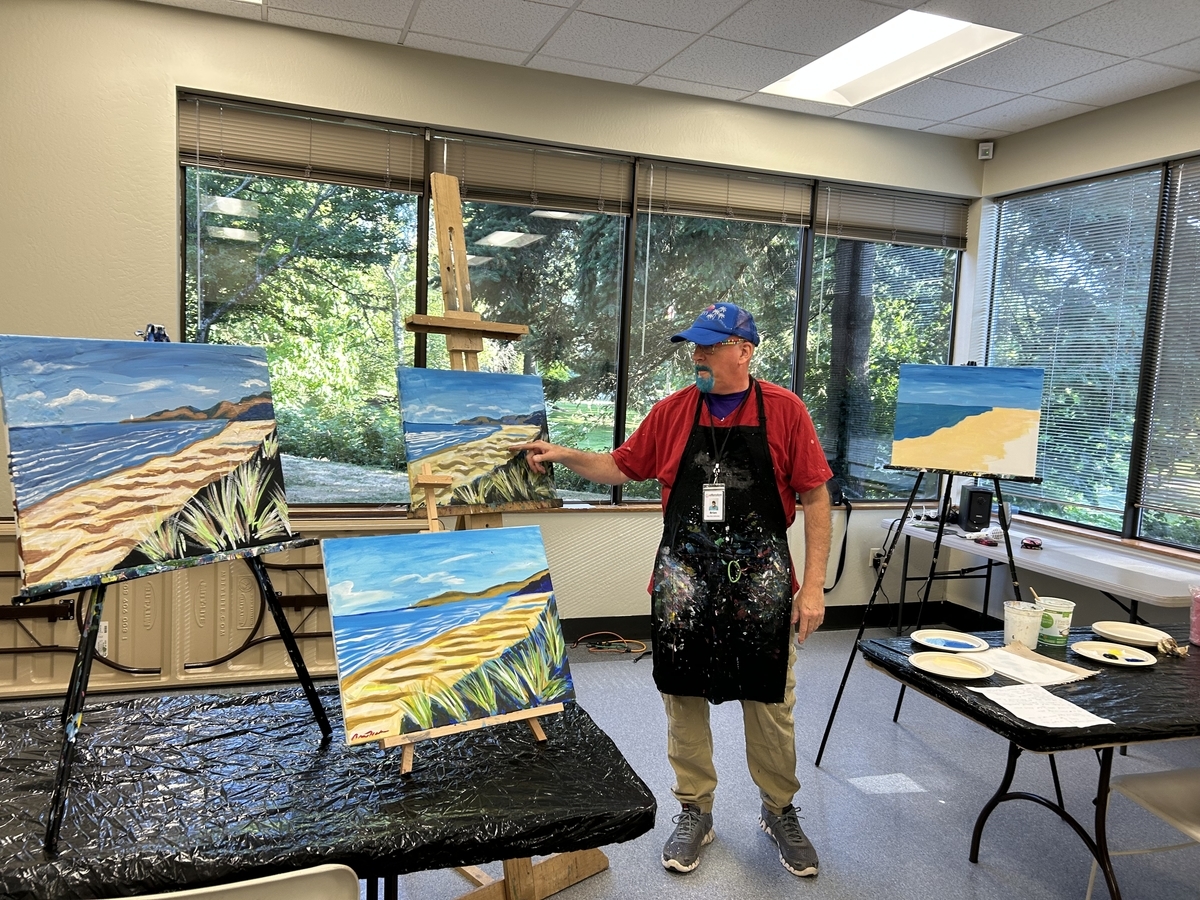 An adult with a paint smeared apron points at a canvas with a beach landscape scene.