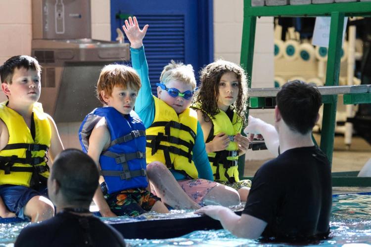 Children in lifejackets sit on the edge of a pool listening to instructor
