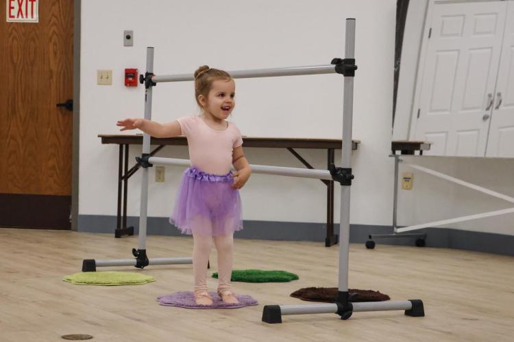 A child smiles with their arm extended in front of a ballet bar, wearing a purple tutu.
