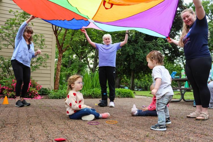 A group of adults hold a multi-color parachute in the air, above a group of toddlers smiling on the ground.