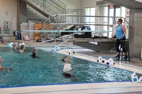 An adult stands on the pool deck instructing those in the water to do exercises.