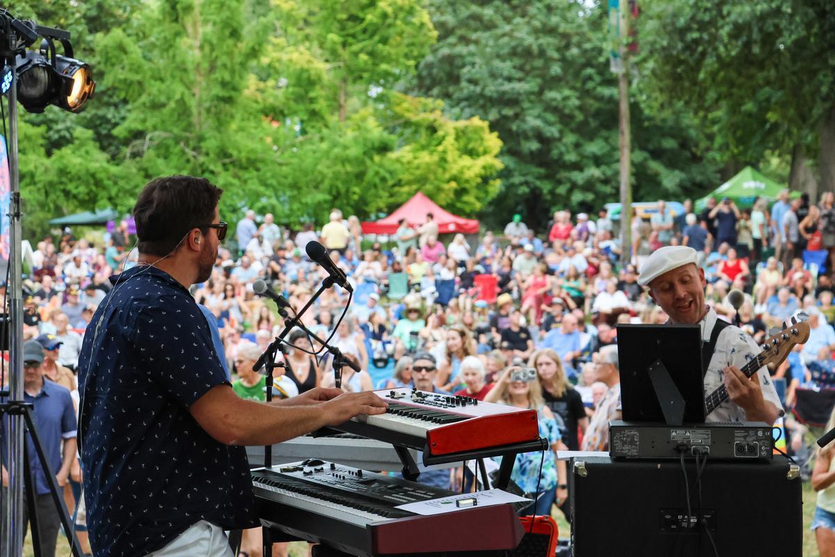 Adults stand on stage of an outdoor concert, playing instruments.