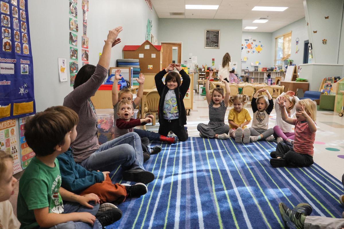 A group of children sit in a circle with their hands over their heads singing a song.