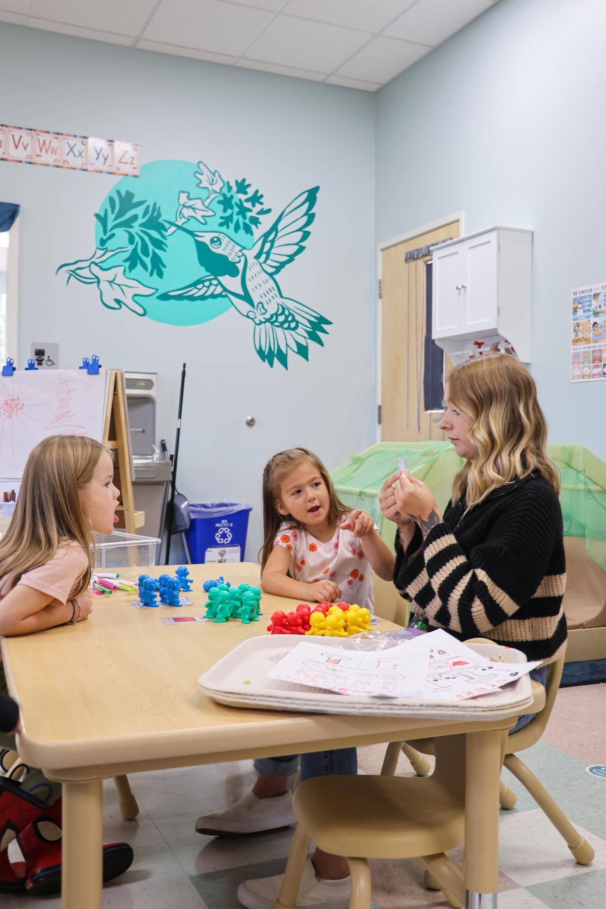 Two children sit at a table with a teacher, pointing at her.