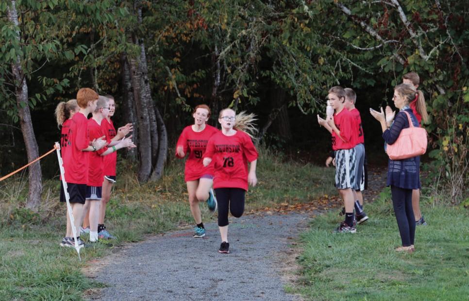 A group of kids in red jerseys run on a gravel trail outside through the trees.