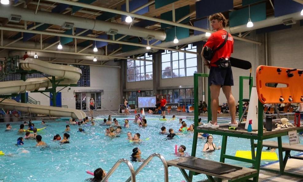 A lifeguard stands high next to a pool filled with swimmers.