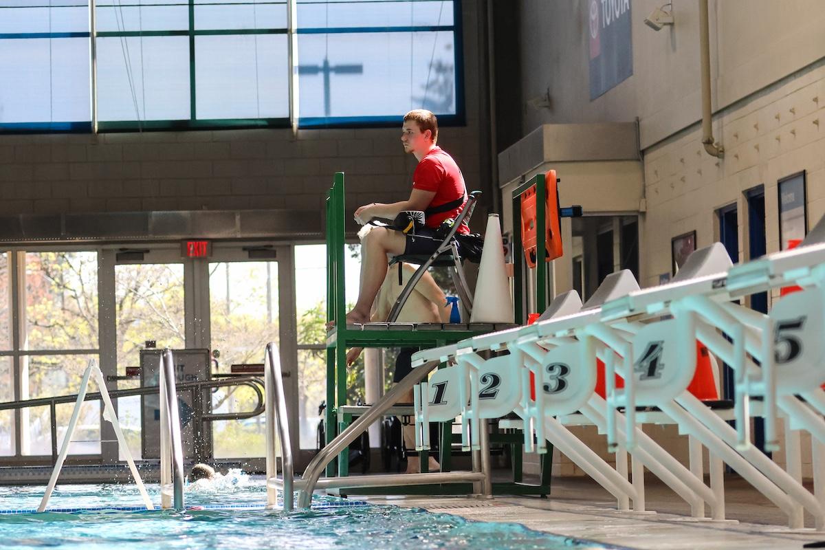An adult in a red shirt sits above the pool watching swimmers.