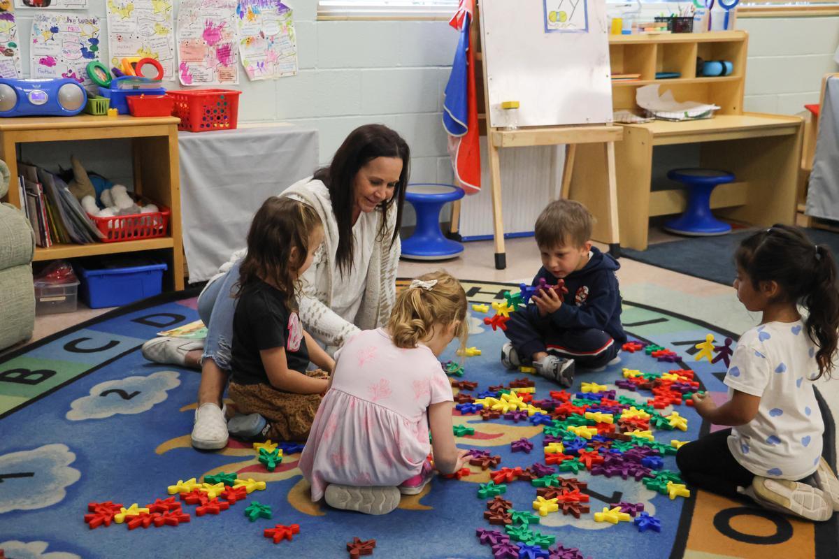 Four children sit on the floor playing with toys alongside their teacher.
