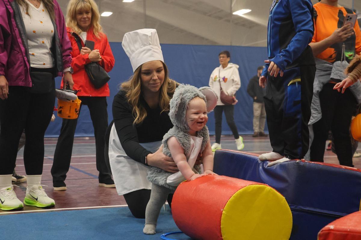 A parent dressed like a chef stands with their child, dressed in a mouse costume, standing by a tumbling course.