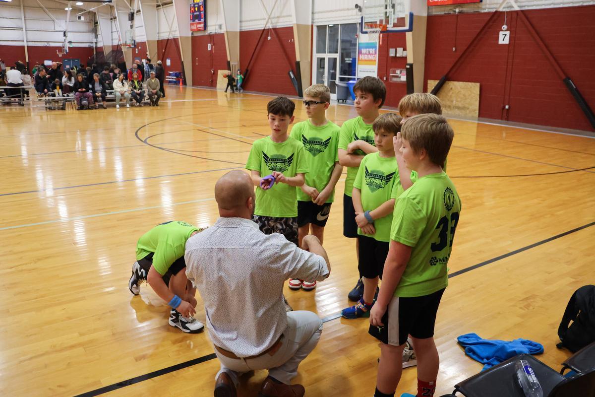 A coach crouches on the ground in front of a team of youth basketball players, pointing and explaining strategy for the second part of the game.