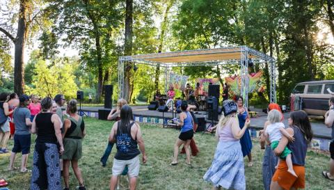 A group of people sit and stand on green grass in front of a large stage with a band playing music.
